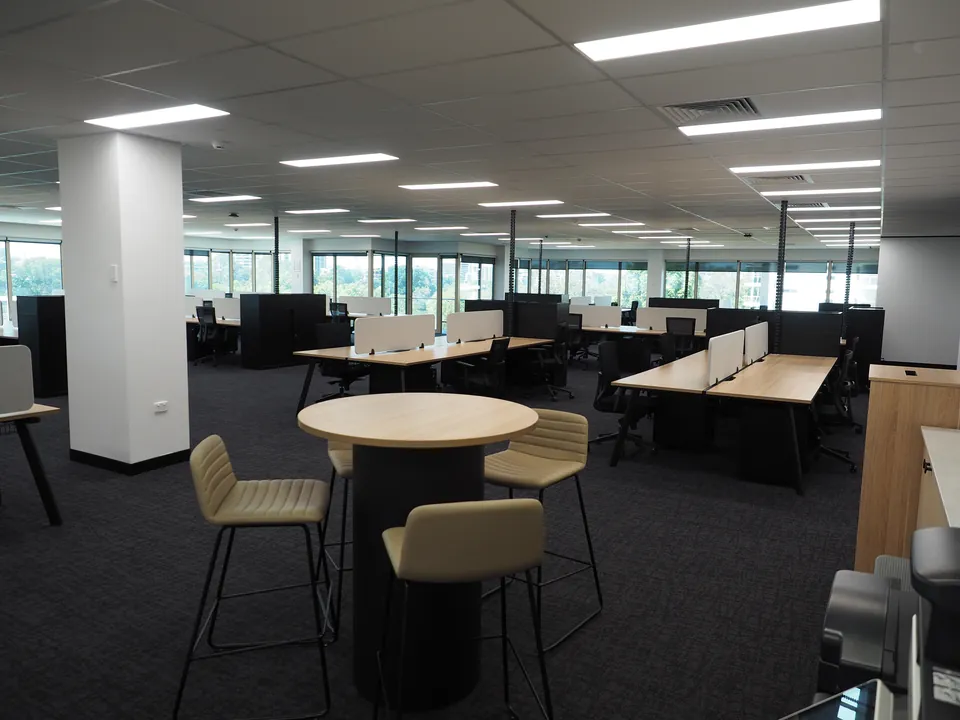 Open-plan office floor with rows of workstations, white desk screens and a round bar-height table with beige stools in the foreground