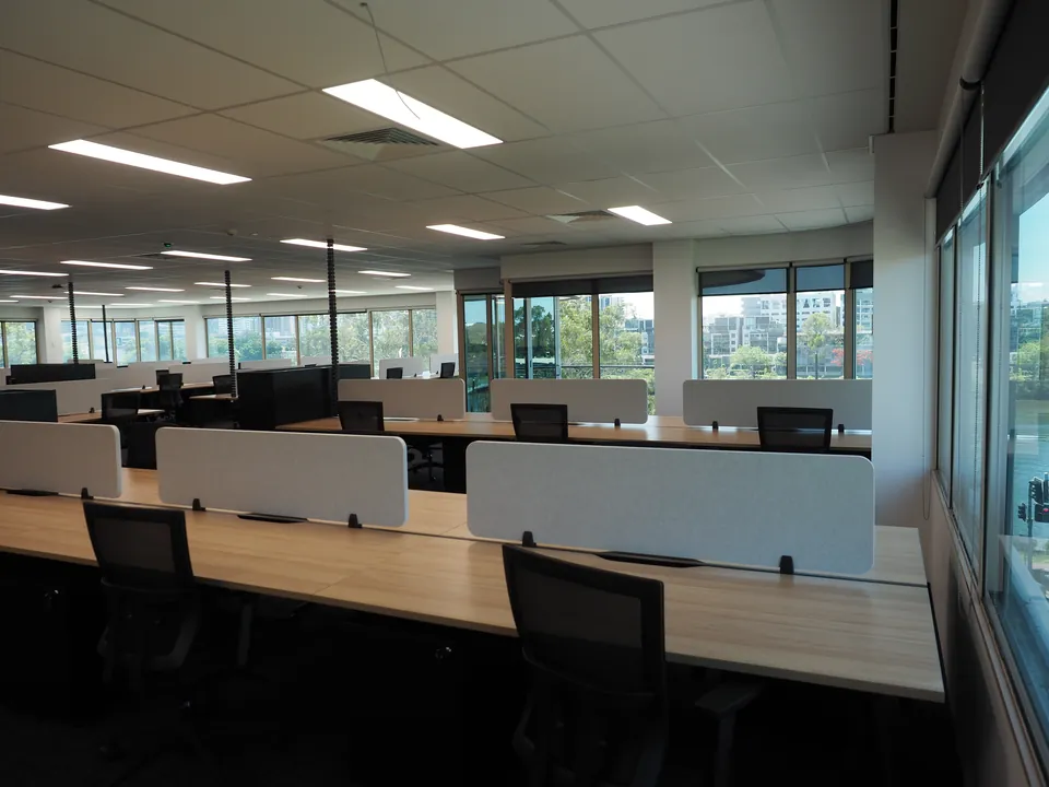 Timber-look workstations with light grey acoustic desk screens and black mesh chairs beside river-view windows