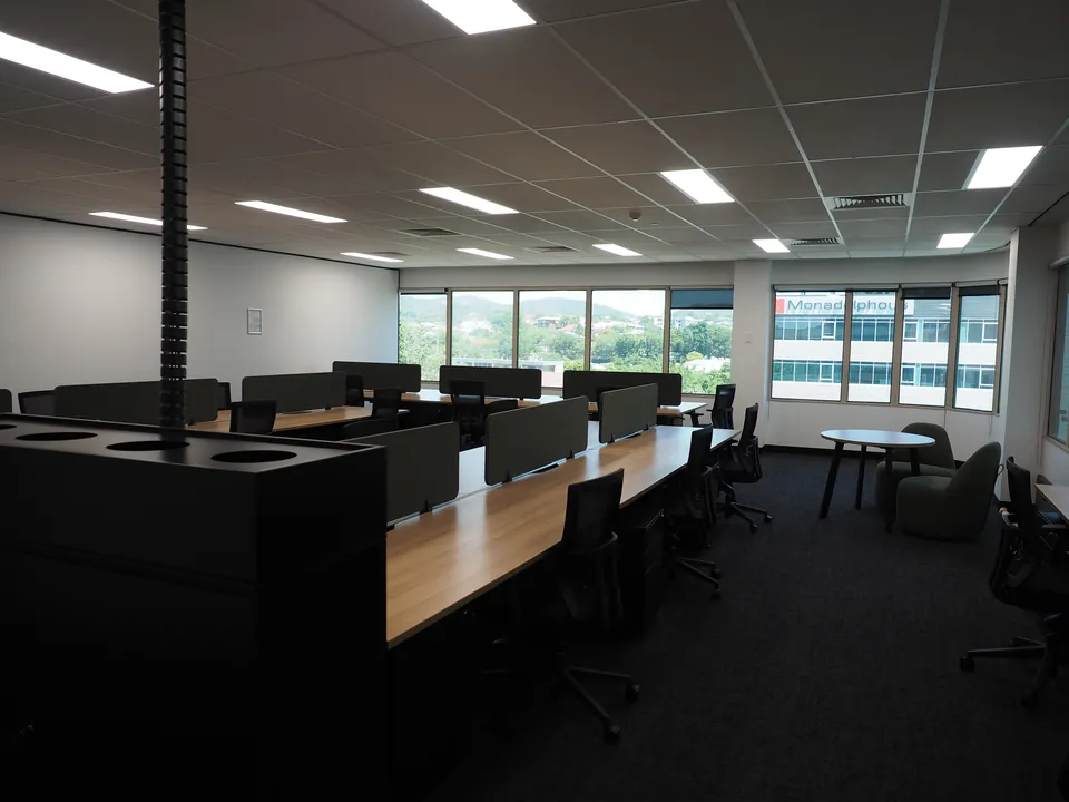Corner of the office with dark timber workstations, green desk screens, a planter-top tambour and green tub chairs at a round table