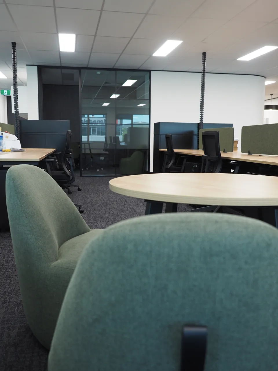 Green upholstered tub chairs around a round timber-look café table, with workstations and green screens behind