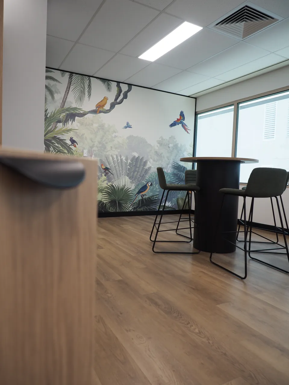 Breakout room with a jungle mural, a round bar-height table on a black drum base and green upholstered stools